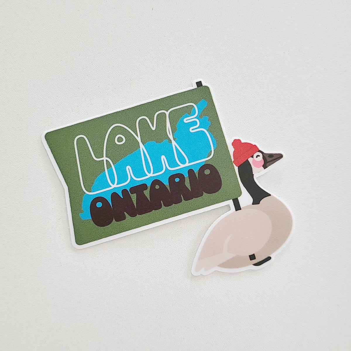 Cute sticker featuring a Canadian goose in a red toque holding a flag that says Lake Ontario. Made in Canada by Mad Love Creative Co.