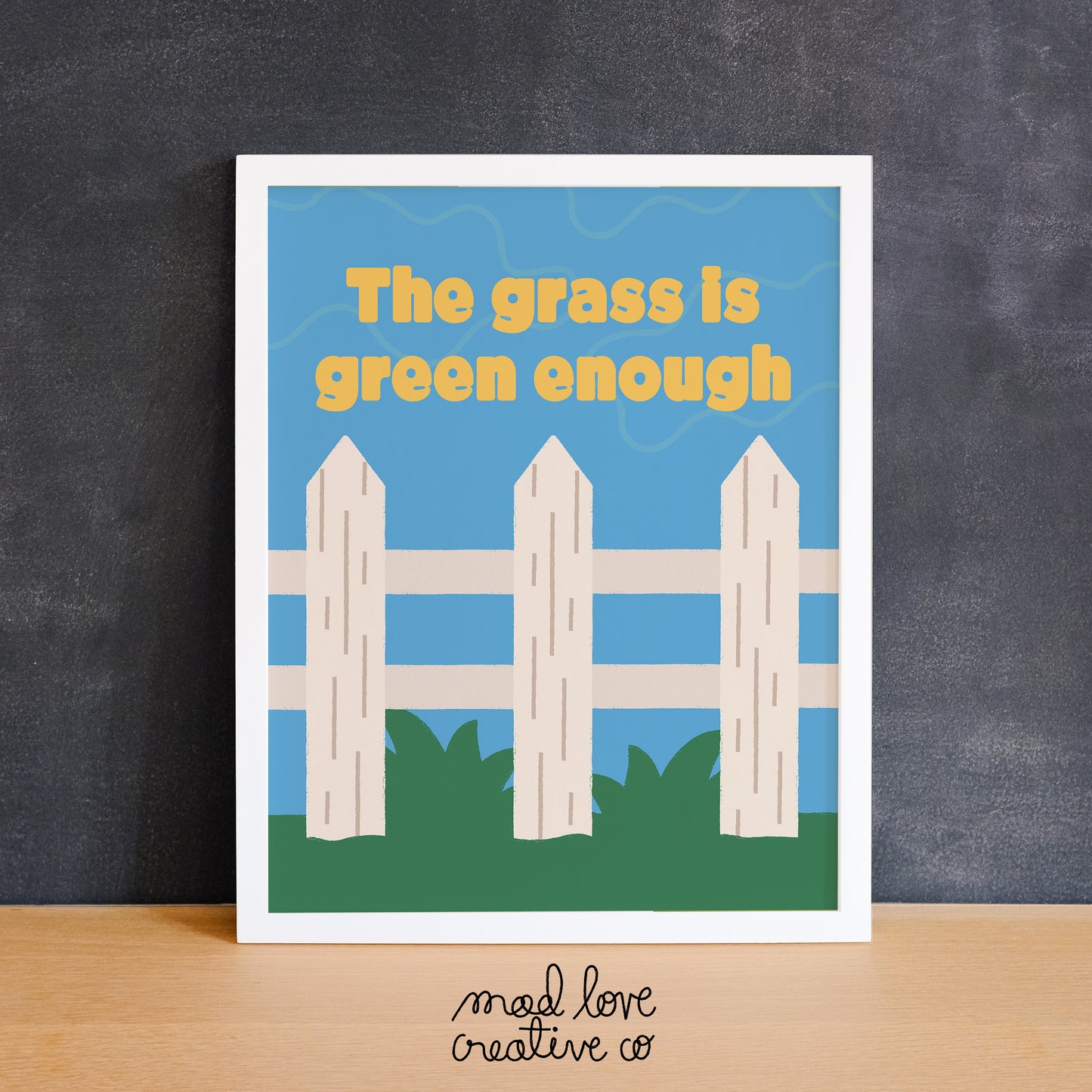 Bright print of a white picket fence on green grass with the phrase "the grass is green enough". Made in Canada by Mad Love Creative Co.