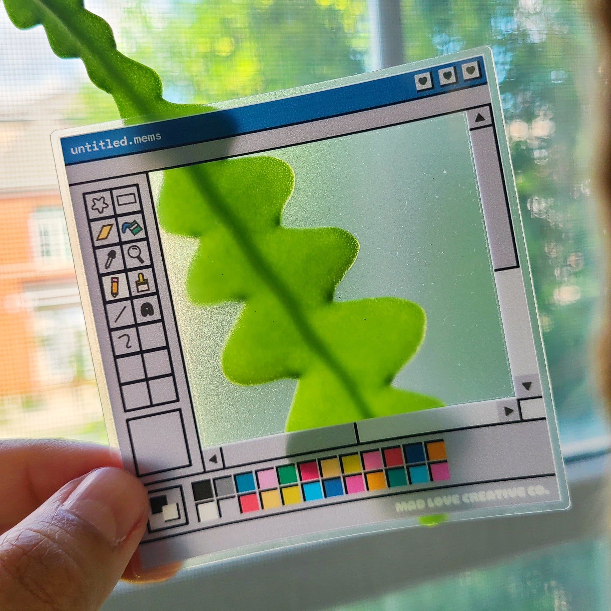 Hand holding a transparent sticker with a digital interface design, showing a leaf shadow on a screen.