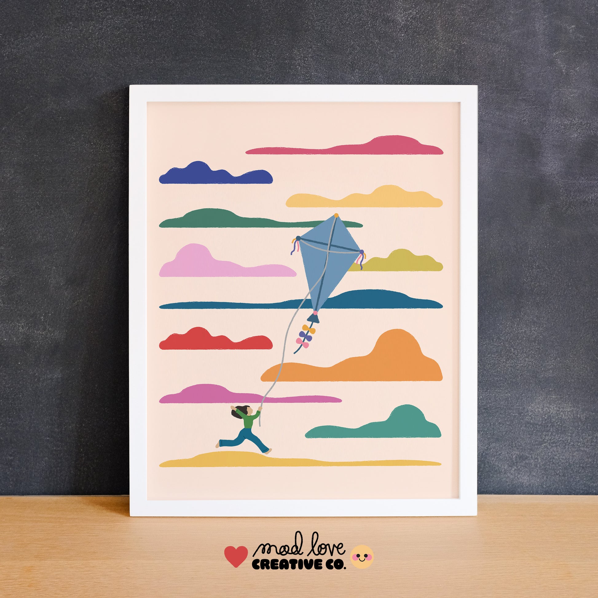 Illustration of a person flying a kite with colorful clouds on a beige background, framed and displayed against a dark wall. Made in Canada by Mad Love Creative Co. 